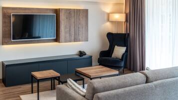 Una sala de estar moderna con un televisor de pantalla plana montado en un panel de madera, un sillón azul oscuro con un cojín, una pequeña mesa de centro de madera y un sofá gris.
