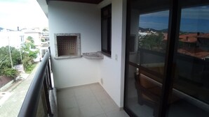 A balcony with a tiled floor, a small window, and a view of buildings and greenery. - Apartamento Praia do Santinho (Florianópolis)