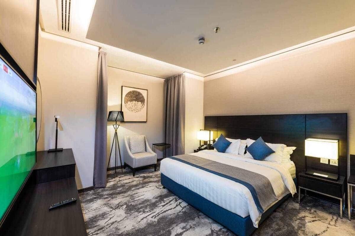 A hotel room with a large bed, a TV on a stand, a desk with a lamp, and a chair.