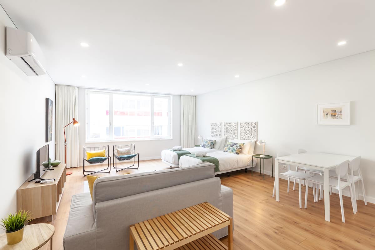 A modern, well-lit room with a sofa, a small wooden table, a bed with a green blanket, a dining table with chairs, and a TV on a wooden stand.