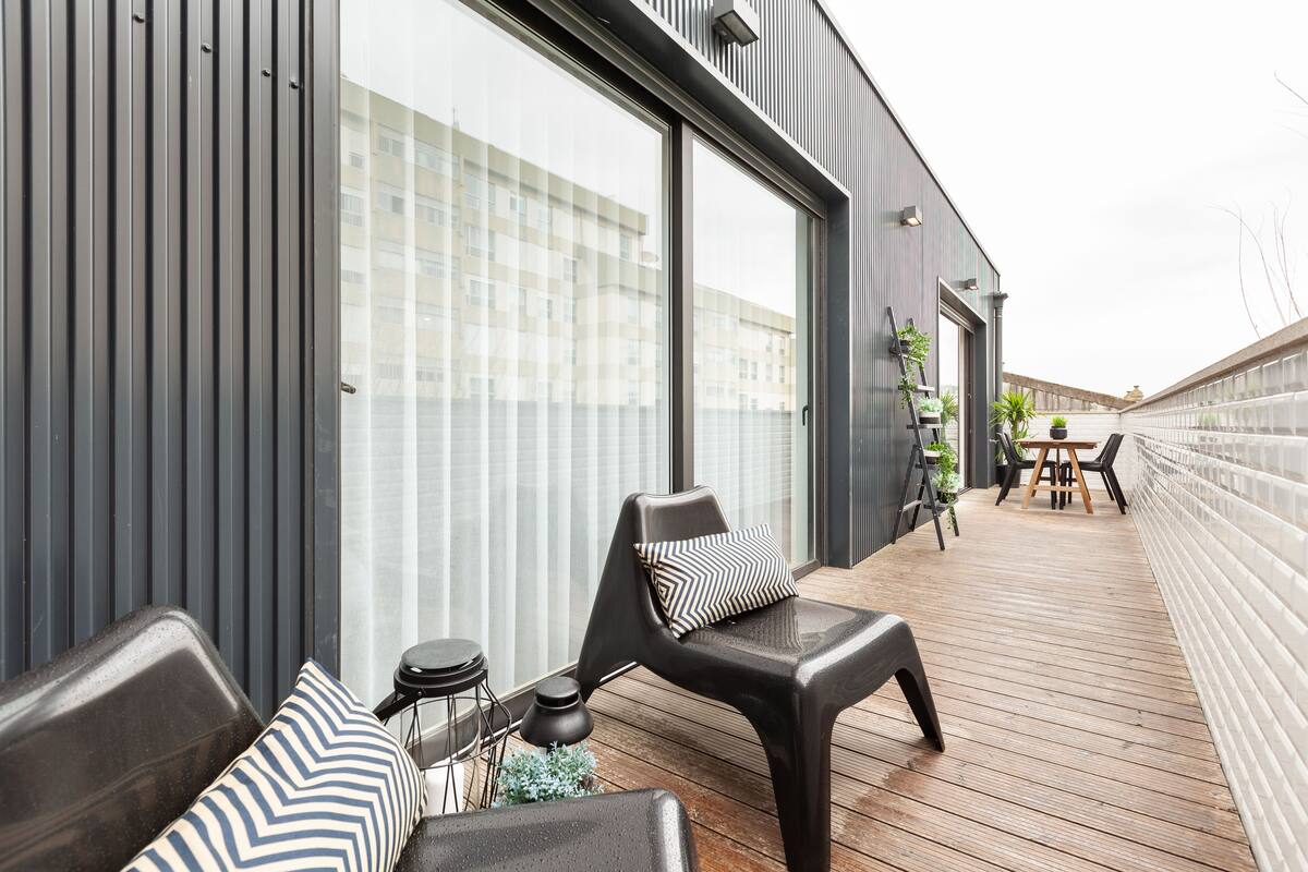 A modern outdoor deck with a wooden floor, a black chair with a striped pillow, a small round table, and a potted plant.