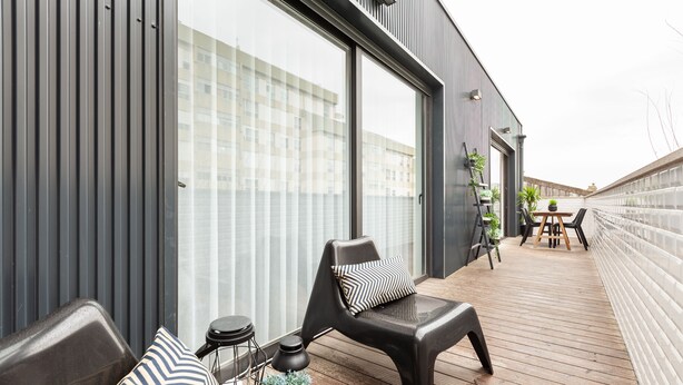 A modern outdoor deck with a wooden floor, a black chair with a striped pillow, a small round table, and a potted plant.
