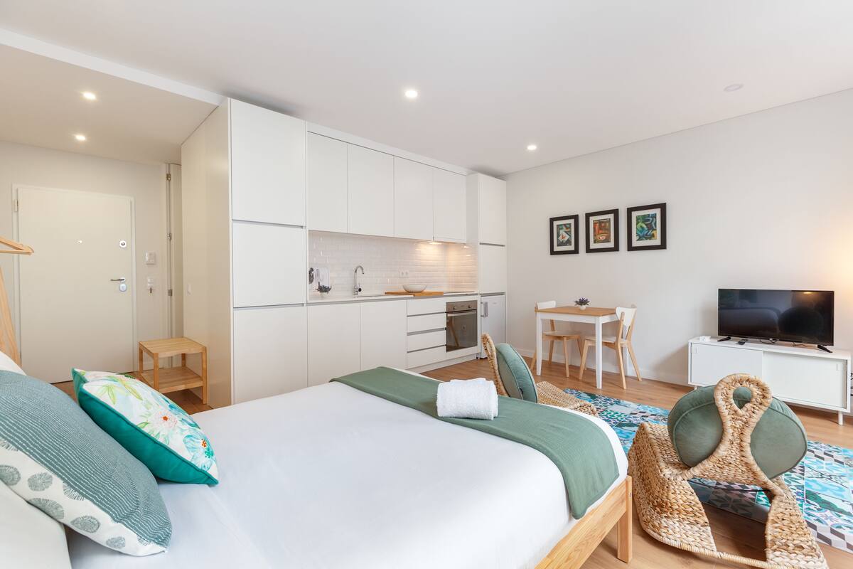 A modern, compact hotel room with a bed, a small kitchenette, a dining area, and a TV.