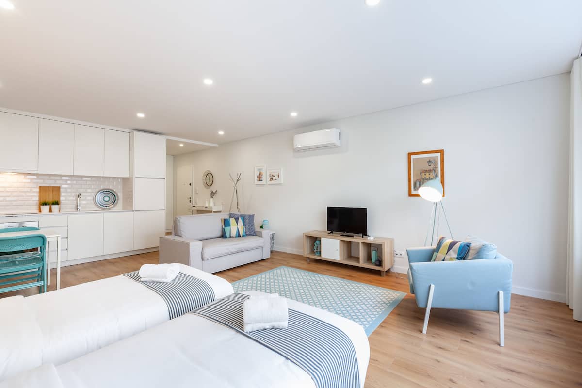 A modern, well-lit room with a large bed, a sofa, a TV, and a small kitchen area.