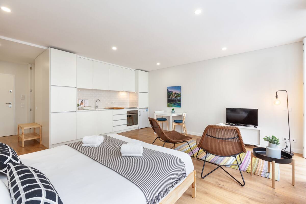 A modern hotel room with a large bed, a desk, a TV, and a kitchenette.