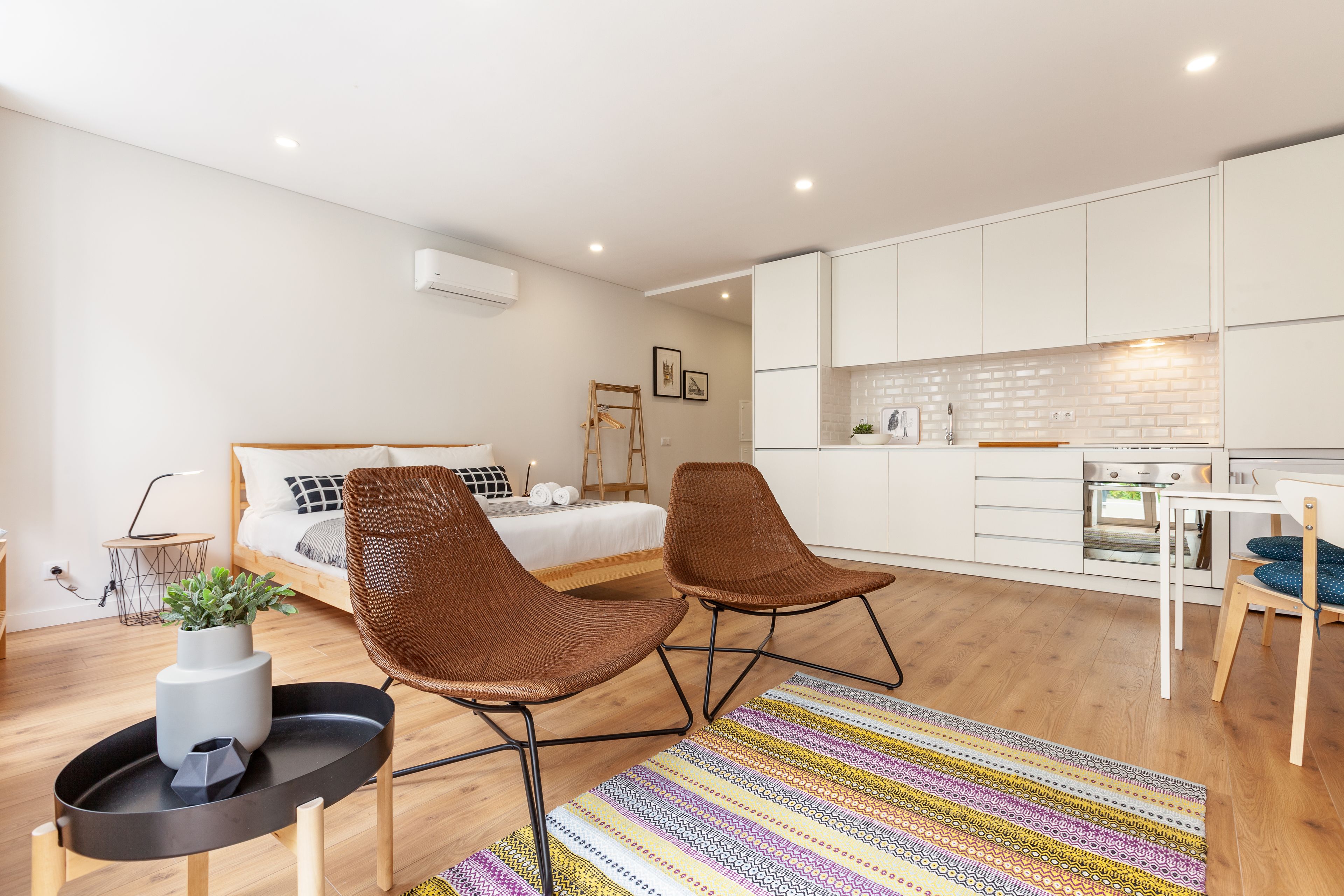 Una habitación de hotel moderna con una cama, dos sillas marrones, una mesita con una planta, una zona de cocina con armarios blancos y una alfombra a rayas.