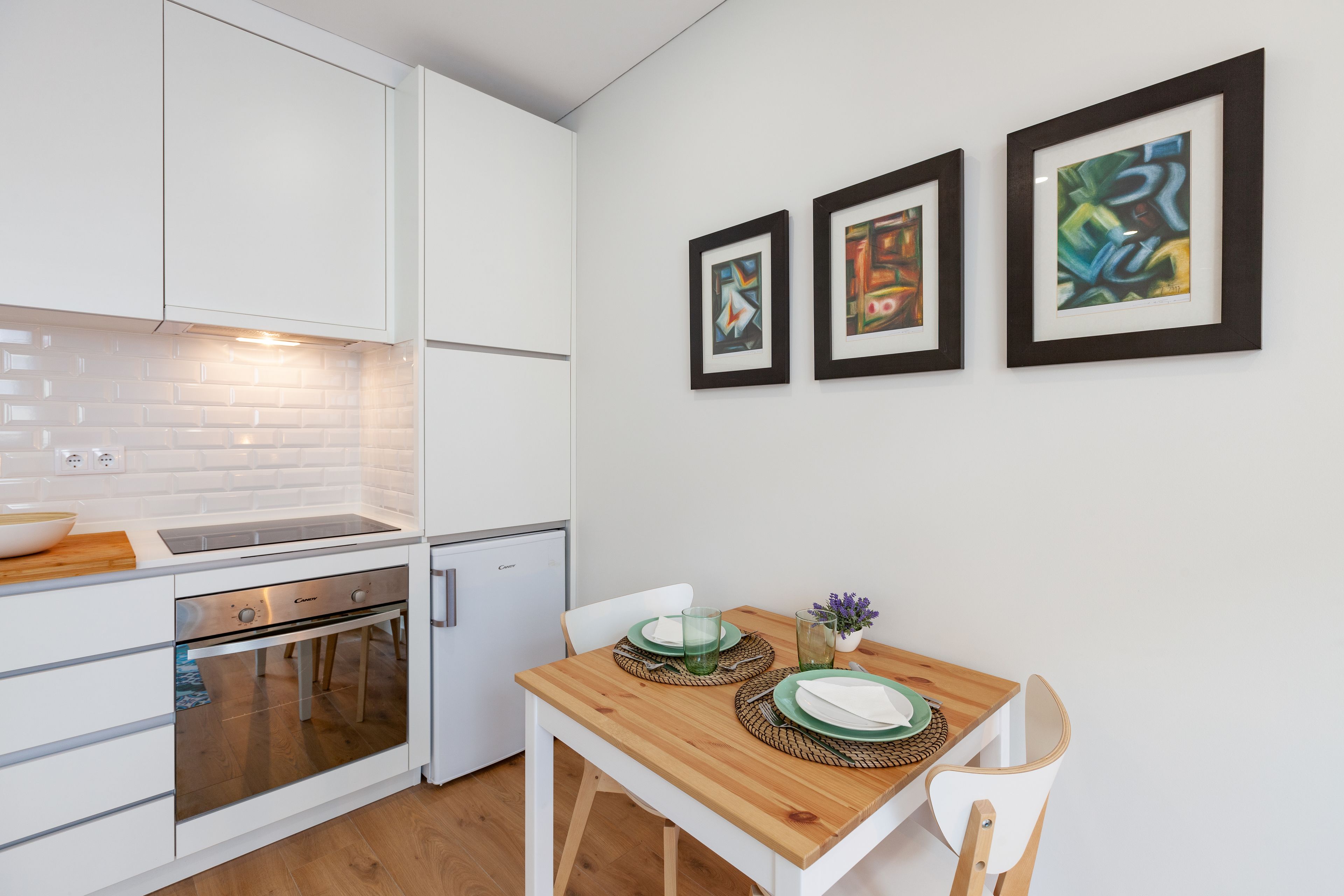 A modern kitchen with white cabinetry, a built-in oven, and a compact refrigerator. There is a wooden dining table with green placemats and a vase with purple flowers. Three framed abstract paintings are hung on the wall above the table.