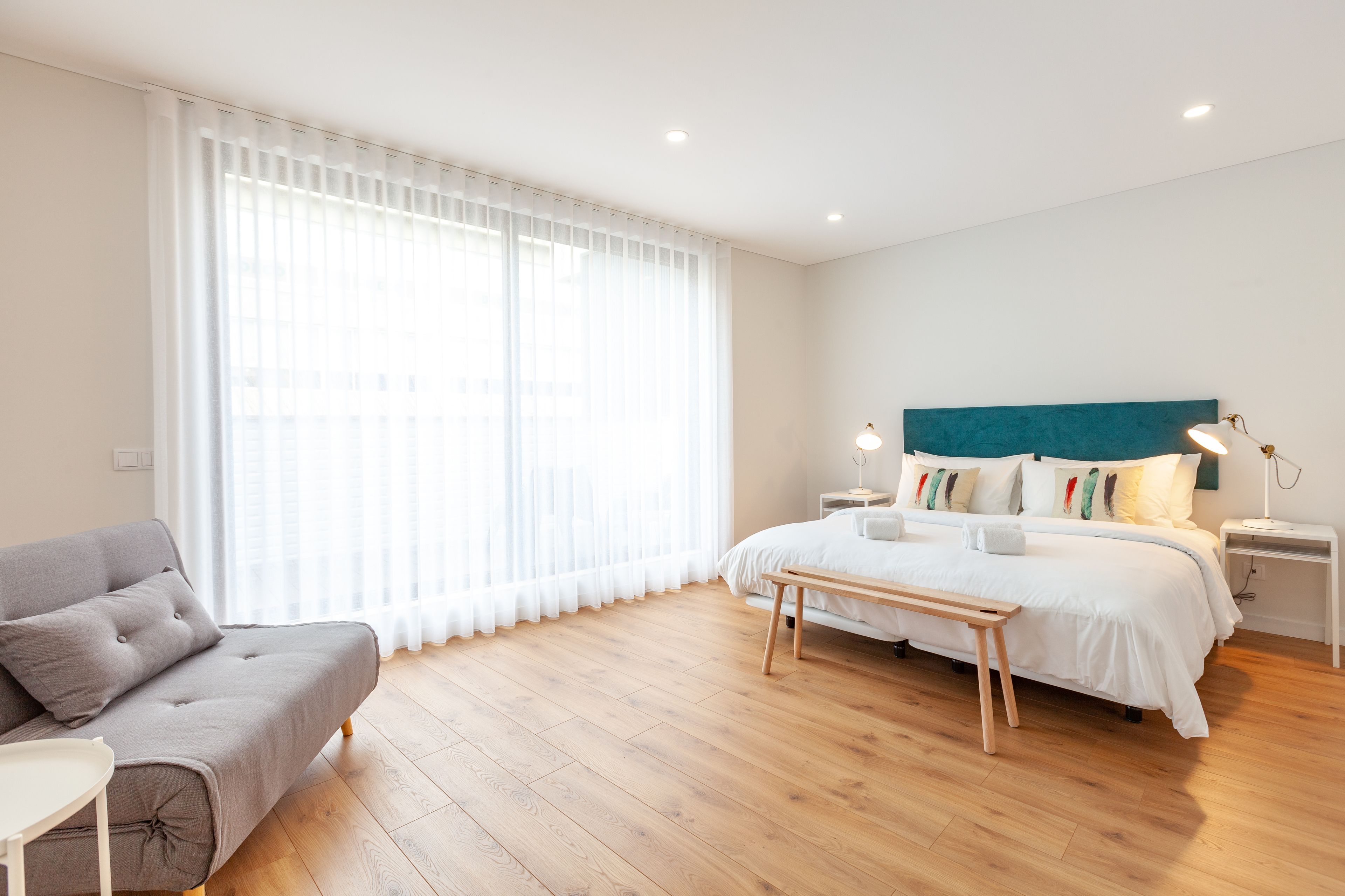 A modern bedroom with a bed, bedside tables, a bench, a sofa, and a large window with sheer curtains.
