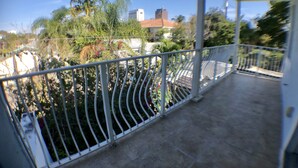 A covered balcony with a curved railing overlooking a garden with various plants and a building in the background.