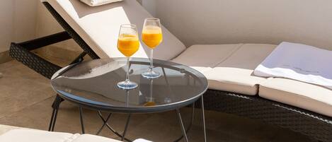 Una terraza en la azotea con una tumbona, una pequeña mesa con dos vasos de zumo de naranja y una vista de una torre histórica al fondo.