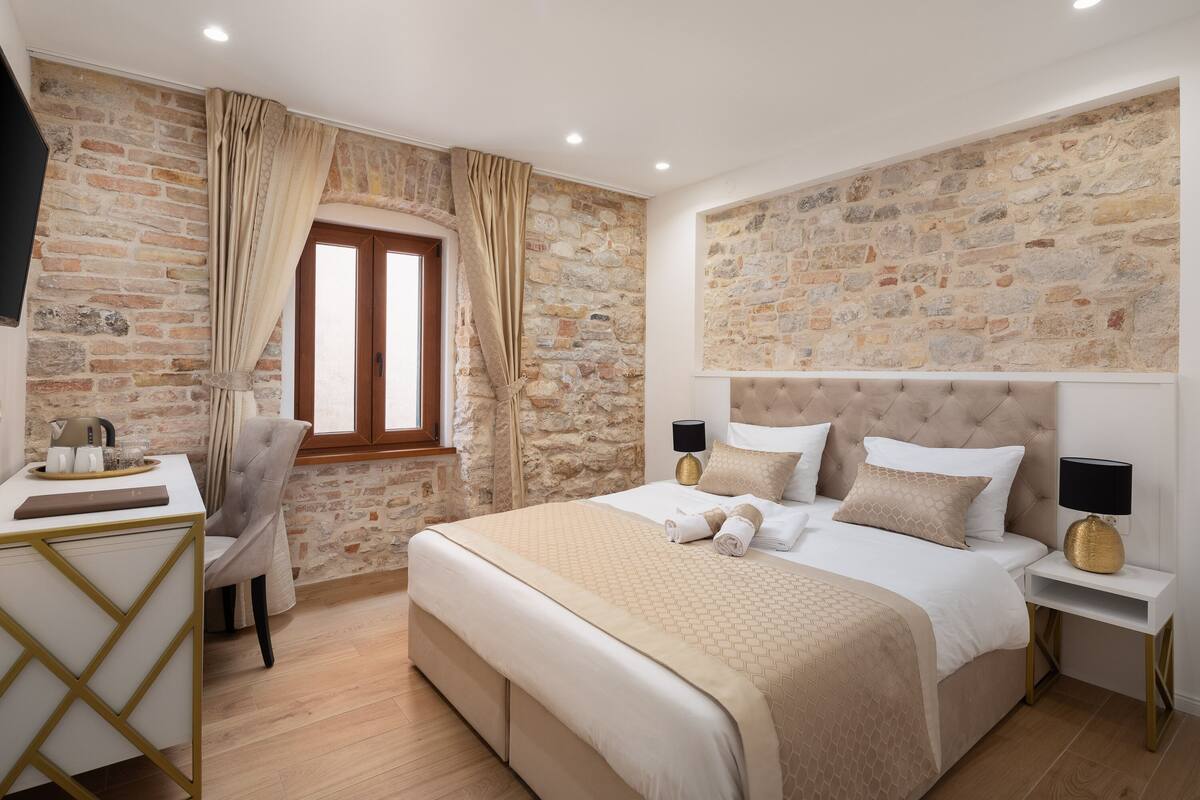 A bedroom with a stone wall, a bed with a beige bedspread, a desk with a chair, and a window with curtains.