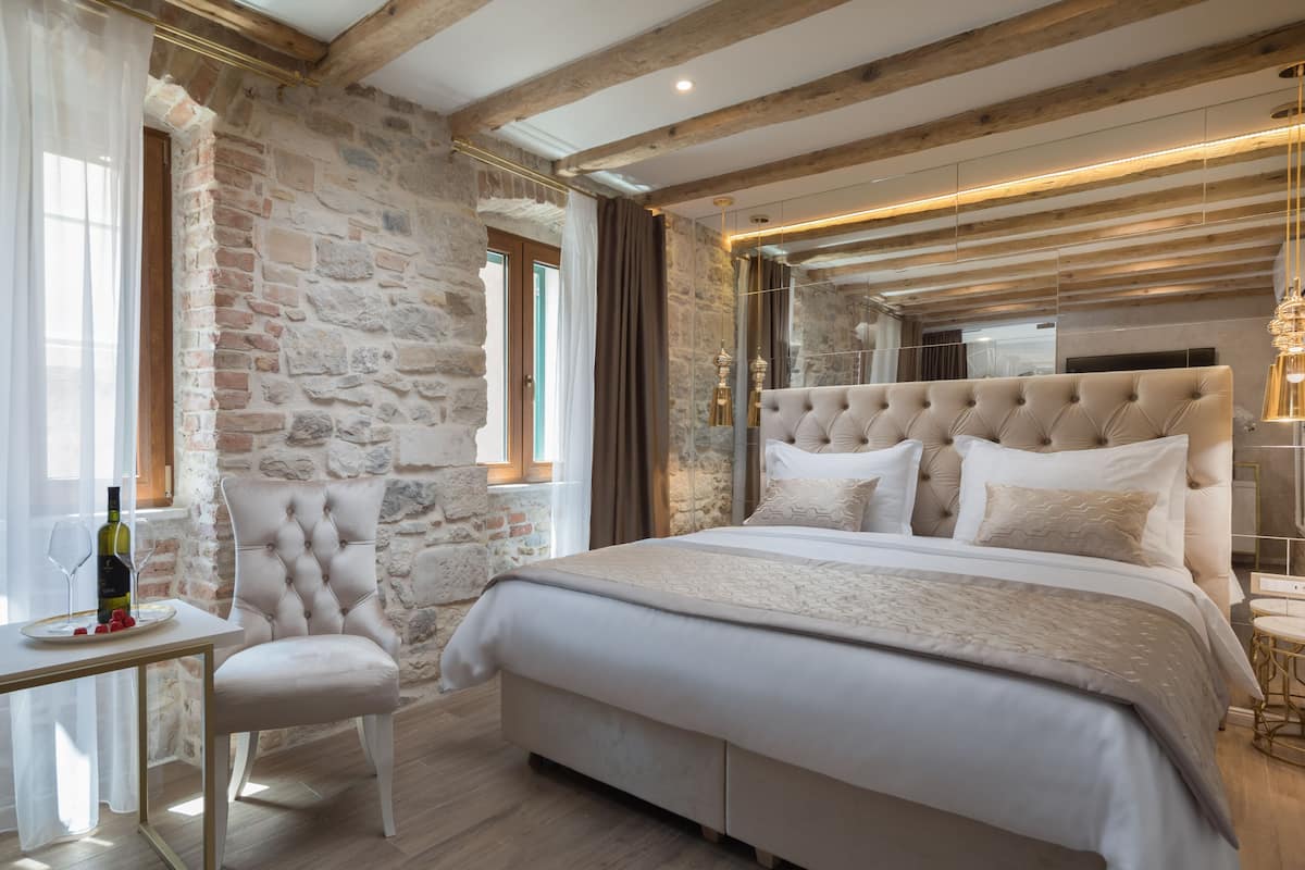 A bedroom with a stone wall, a tufted headboard, a bedside table with a wine bottle, and a view of the outdoors.