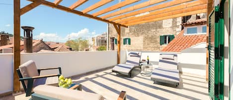 A rooftop terrace with wooden pergola, two lounge chairs, a table with a bottle, and a bowl of fruit.