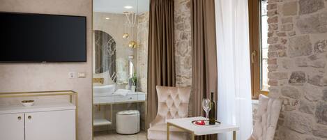 A modern hotel room with a stone wall, a flat-screen TV, a white cabinet, a tufted armchair, a small table with a bottle and fruit, and a glass-enclosed bathroom.