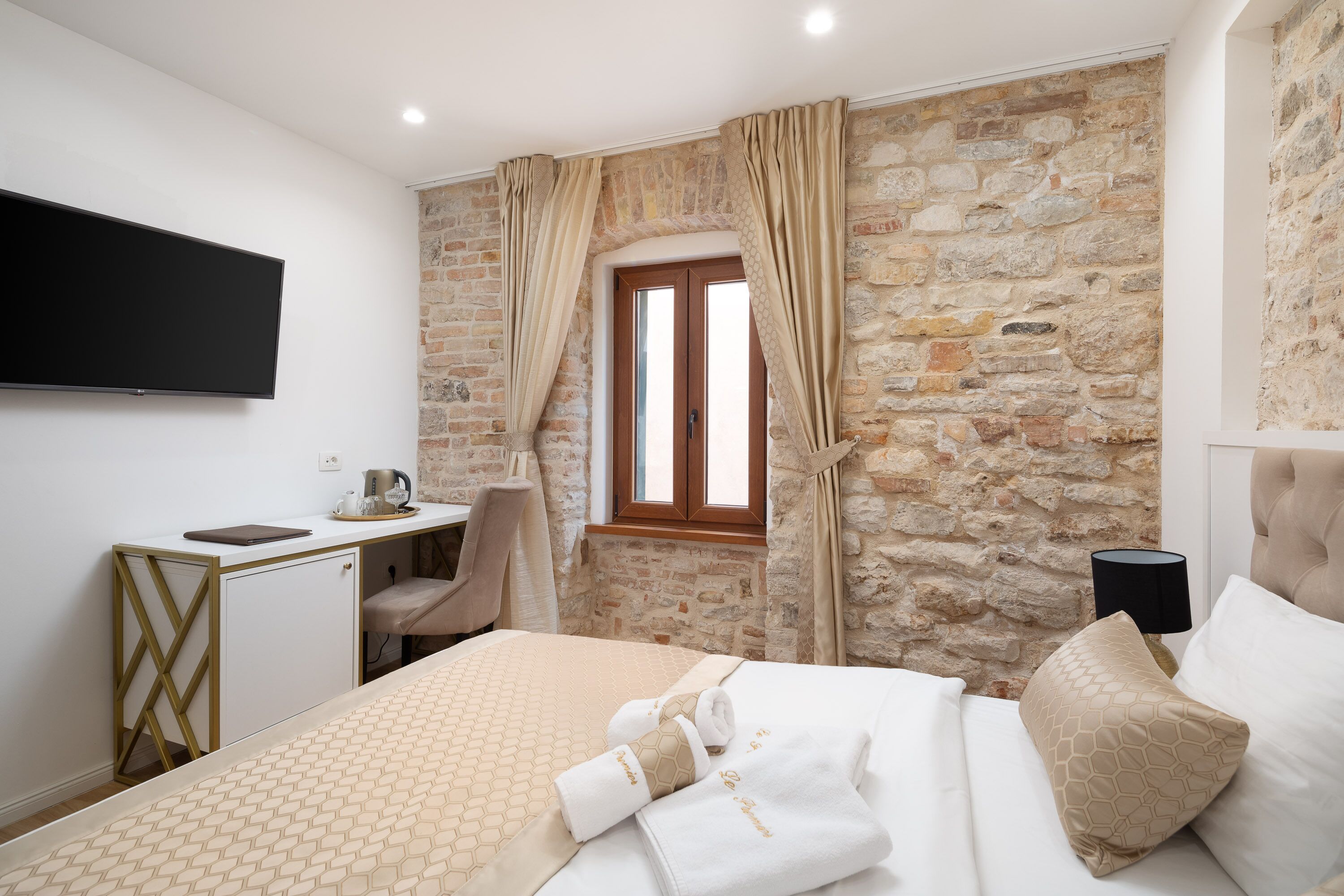 Habitación de hotel con pared de piedra, cama con colcha estampada, escritorio con silla, televisor de pantalla plana y ventana con cortinas.