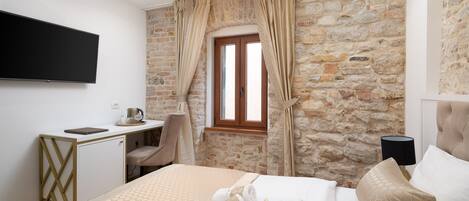 Habitación de hotel con pared de piedra, cama con colcha estampada, escritorio con silla, televisor de pantalla plana y ventana con cortinas.