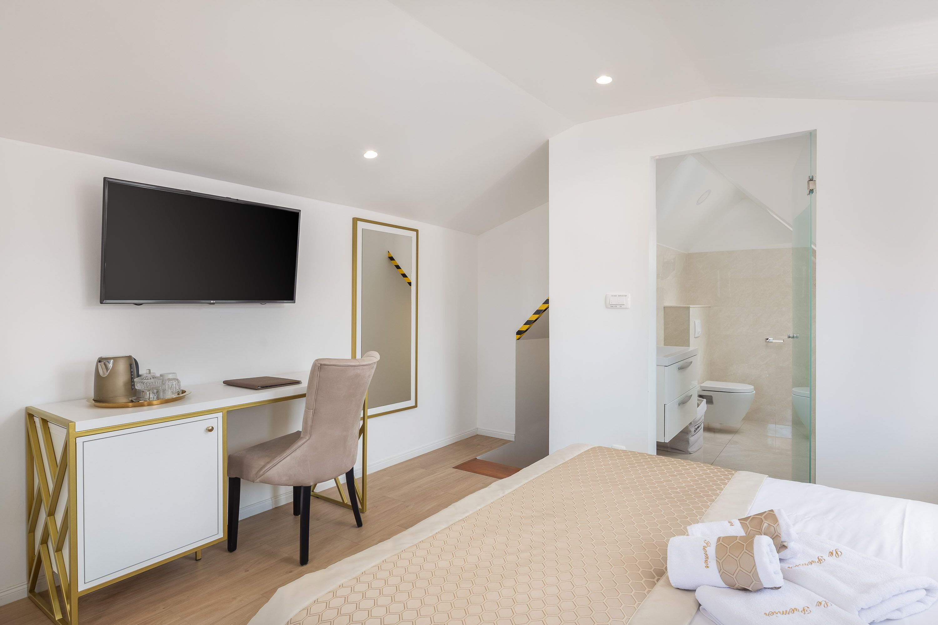 A modern hotel room with a bed, a desk with a TV, a chair, a mirror, and a bathroom with a glass shower.