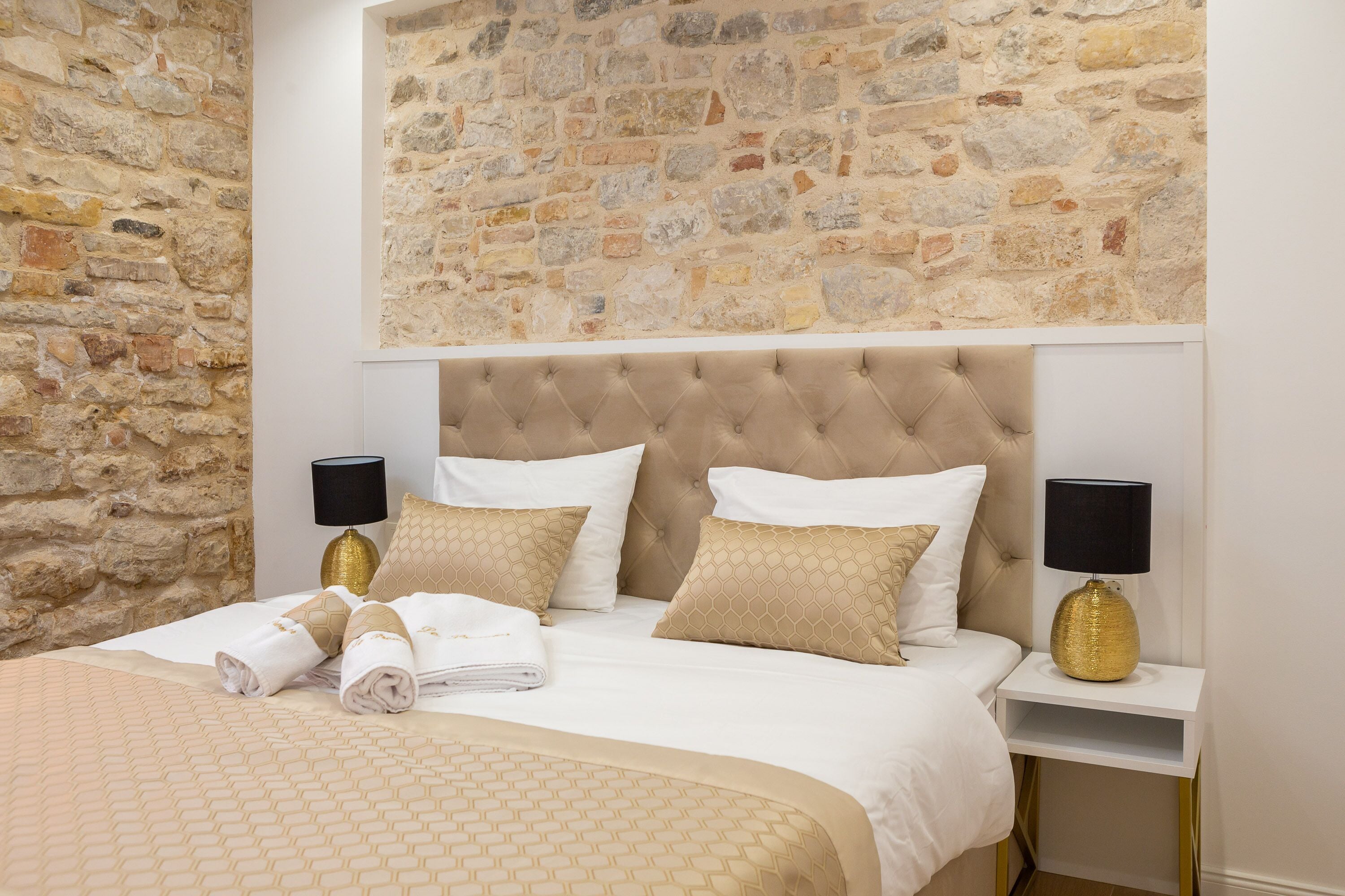 A bedroom with a stone wall, a tufted headboard, two bedside tables with lamps, and neatly folded towels.