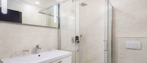 Un baño moderno con ducha envuelta en cristal, un lavamanos blanco con espejo encima y un inodoro con botón de descarga.