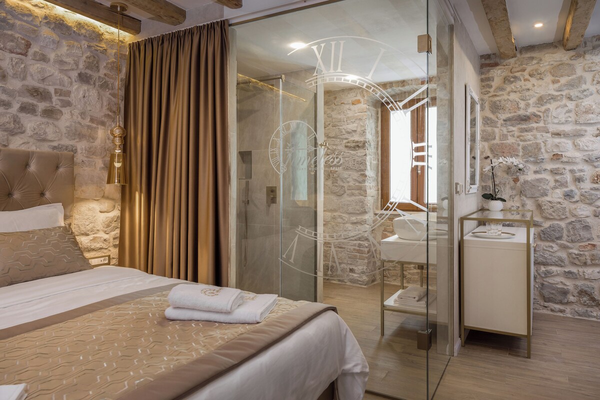 A bedroom with a stone wall, a bed with a beige bedspread, a glass shower enclosure, and a bedside table with a lamp.