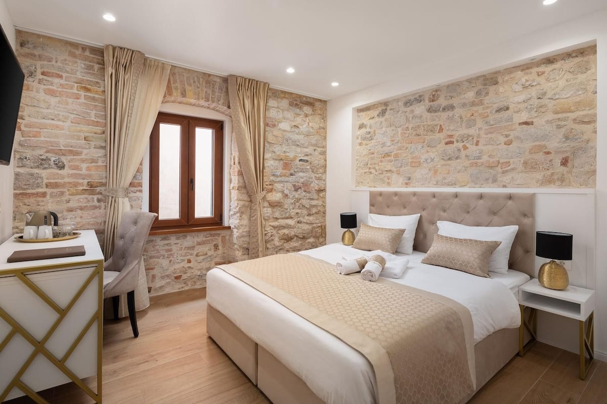 A bedroom with a stone wall, a bed with a beige headboard, a bedside table with a lamp, and a desk with a chair.