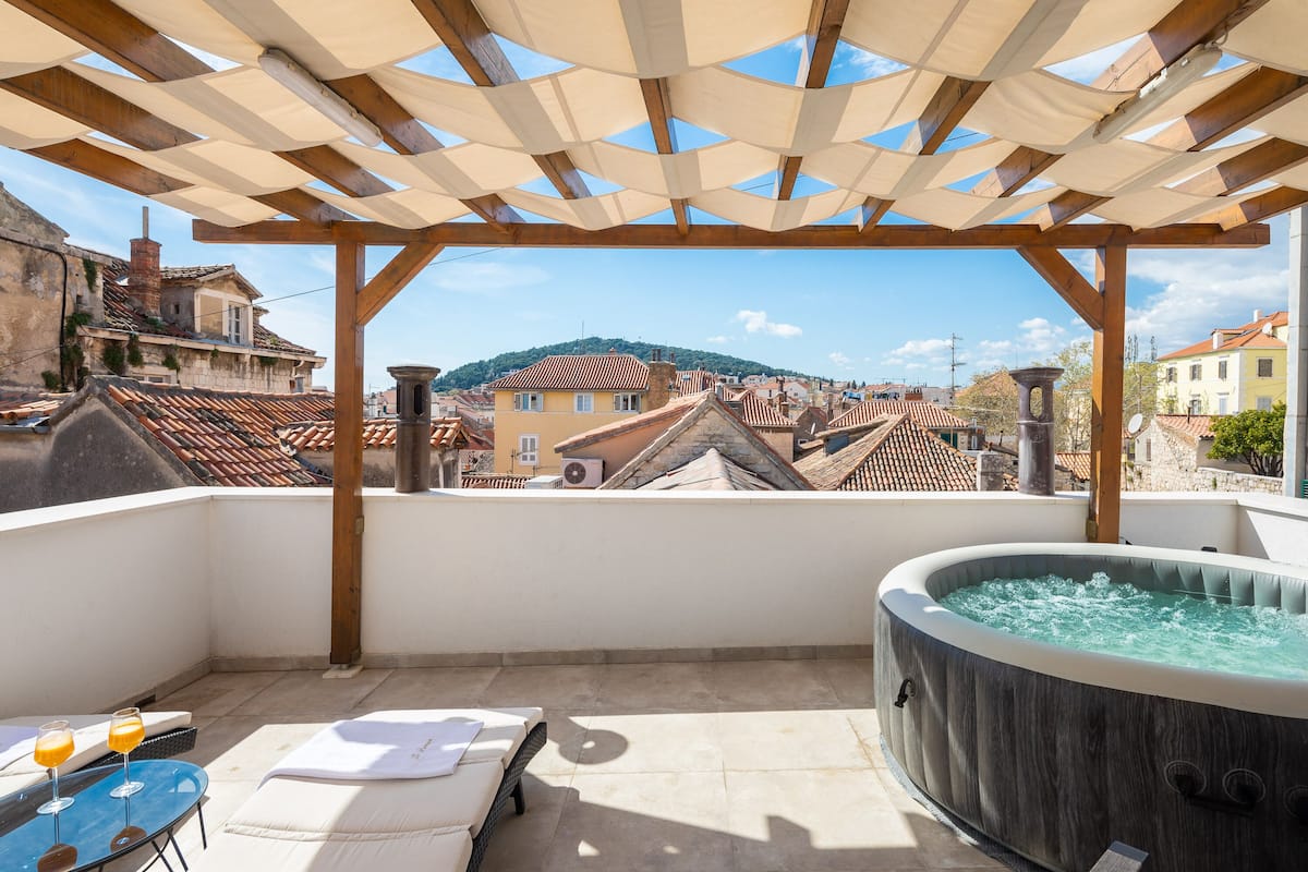 A rooftop terrace with a wooden pergola, a hot tub, lounge chairs, and a view of the cityscape.