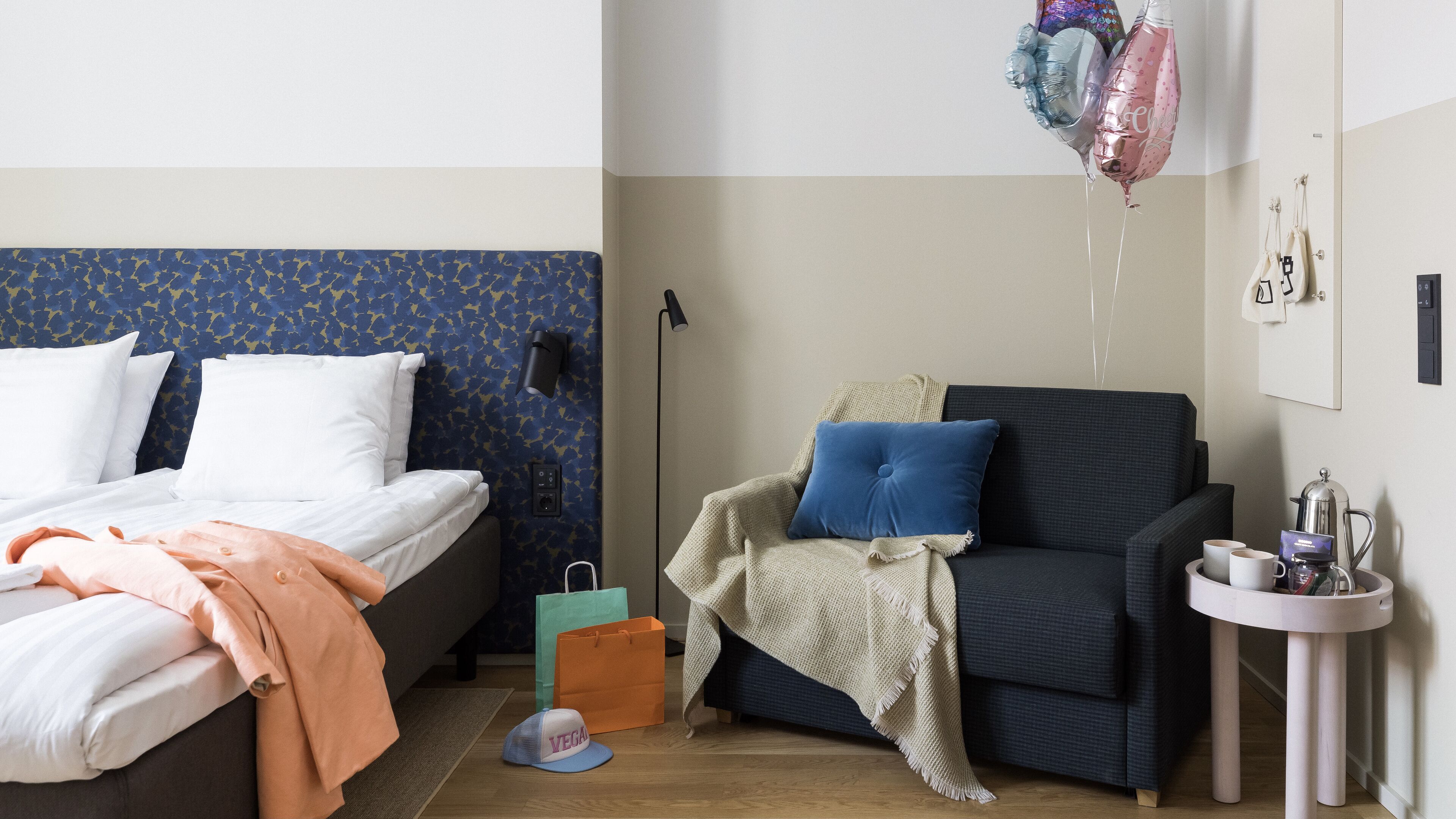 A hotel room with a bed, a sofa, a small table with a coffee pot, and a wall-mounted light.