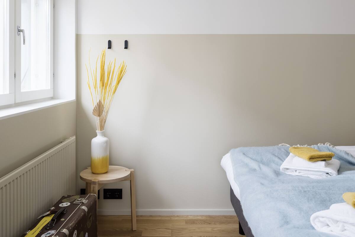 A hotel room with a bed, a small table with a vase of dried plants, a suitcase, and a radiator.