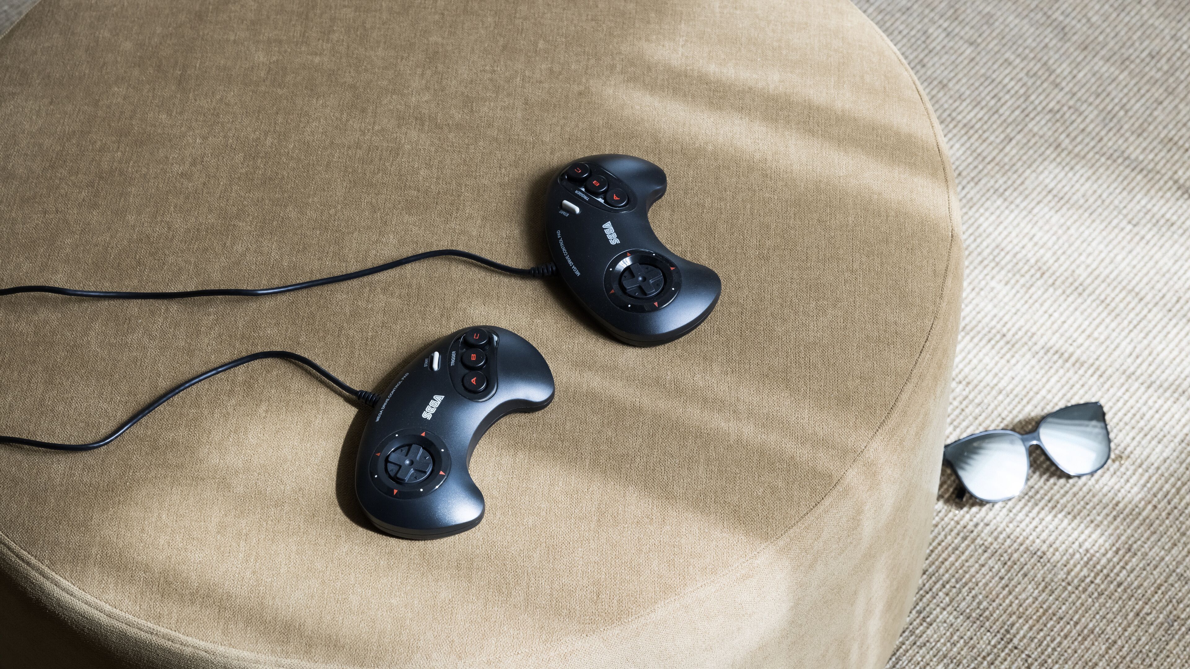 Deux manettes de jeu noires sont posées sur un canapé beige, à côté d’une paire de lunettes de soleil.