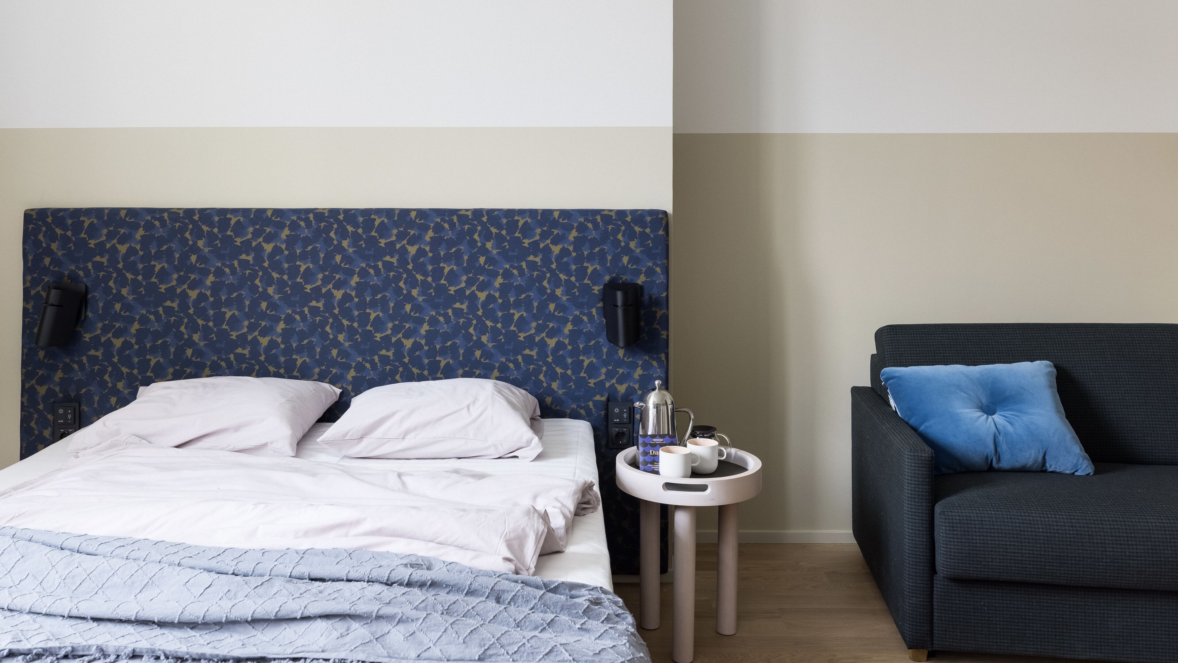 Ein Hotelzimmer mit Bett, einem kleinen Beistelltisch mit Kaffeemaschine und einem dunklen Sofa mit blauem Kissen.