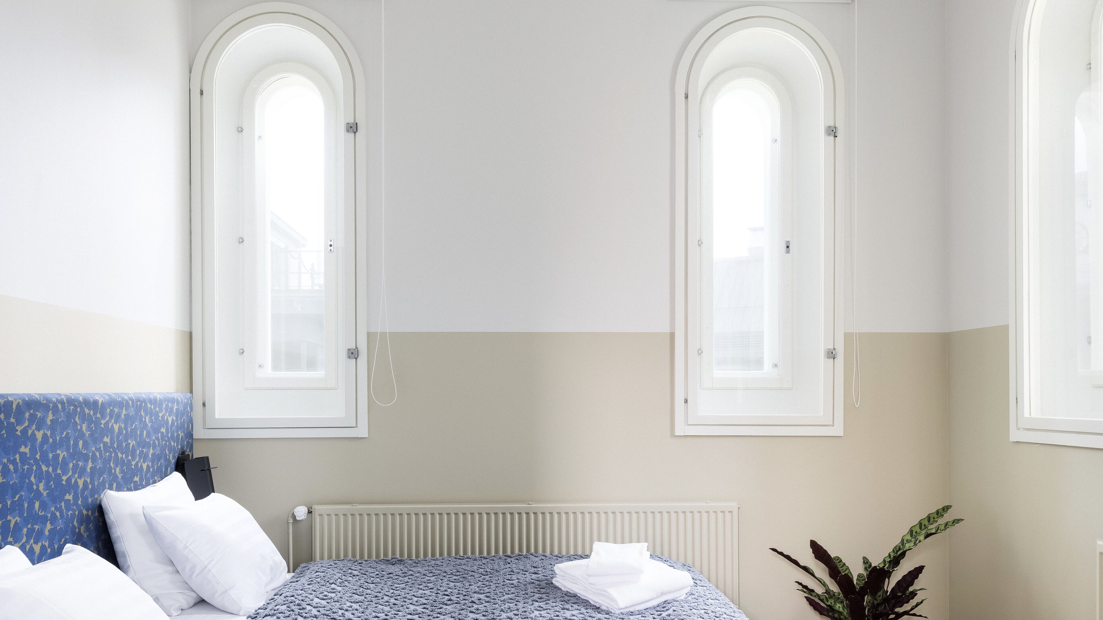 Un dormitorio con una cama, dos ventanas arqueadas, un radiador y una planta.