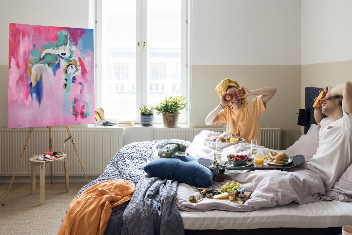 A hotel room with a bed, a painting on the wall, a window with a view, and a breakfast setup on the bed.