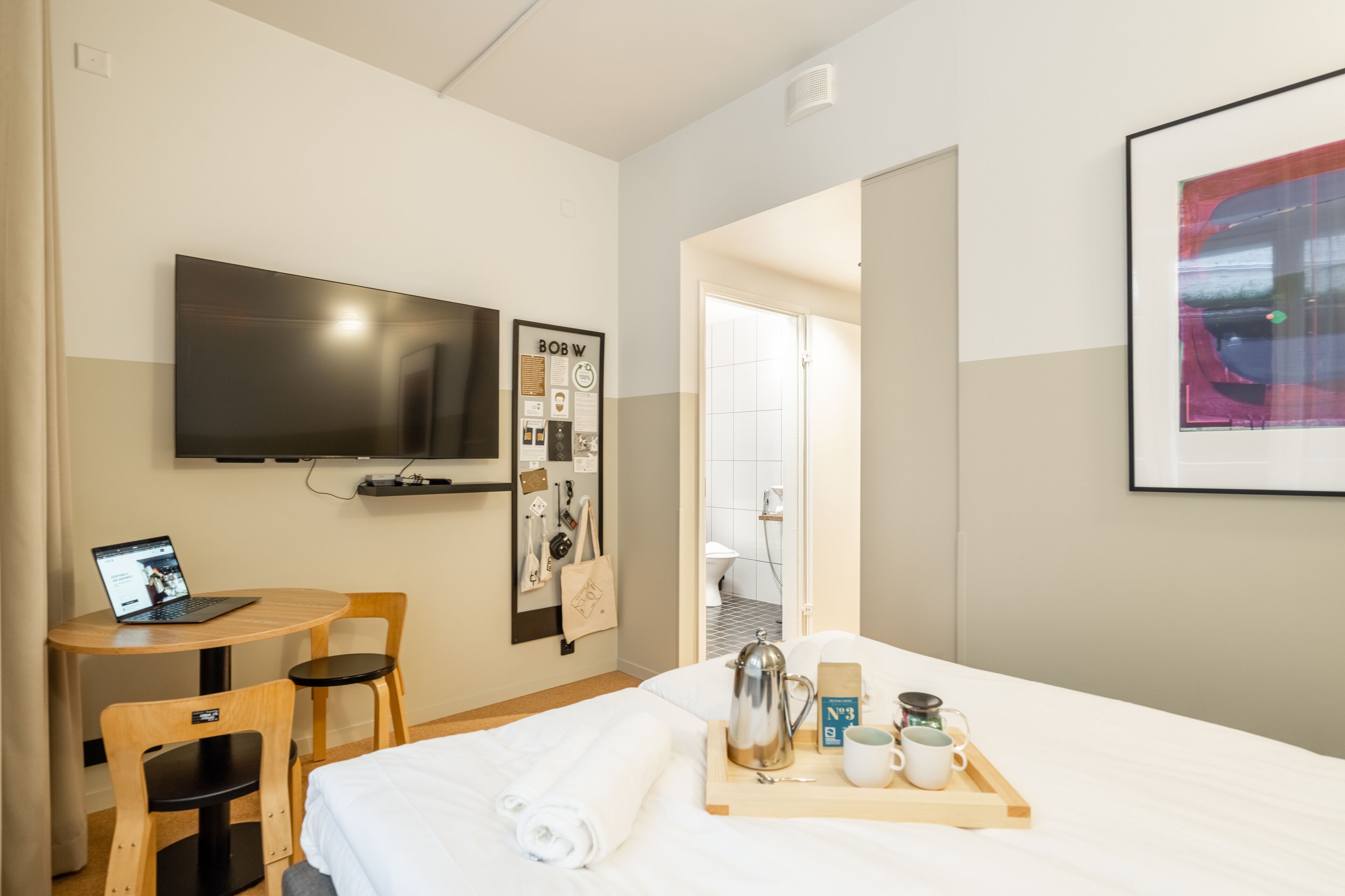 Une chambre d’hôtel avec un lit, un bureau avec un ordinateur portable, un téléviseur fixé au mur, une photo encadrée et un plateau avec une cafetière et des tasses.