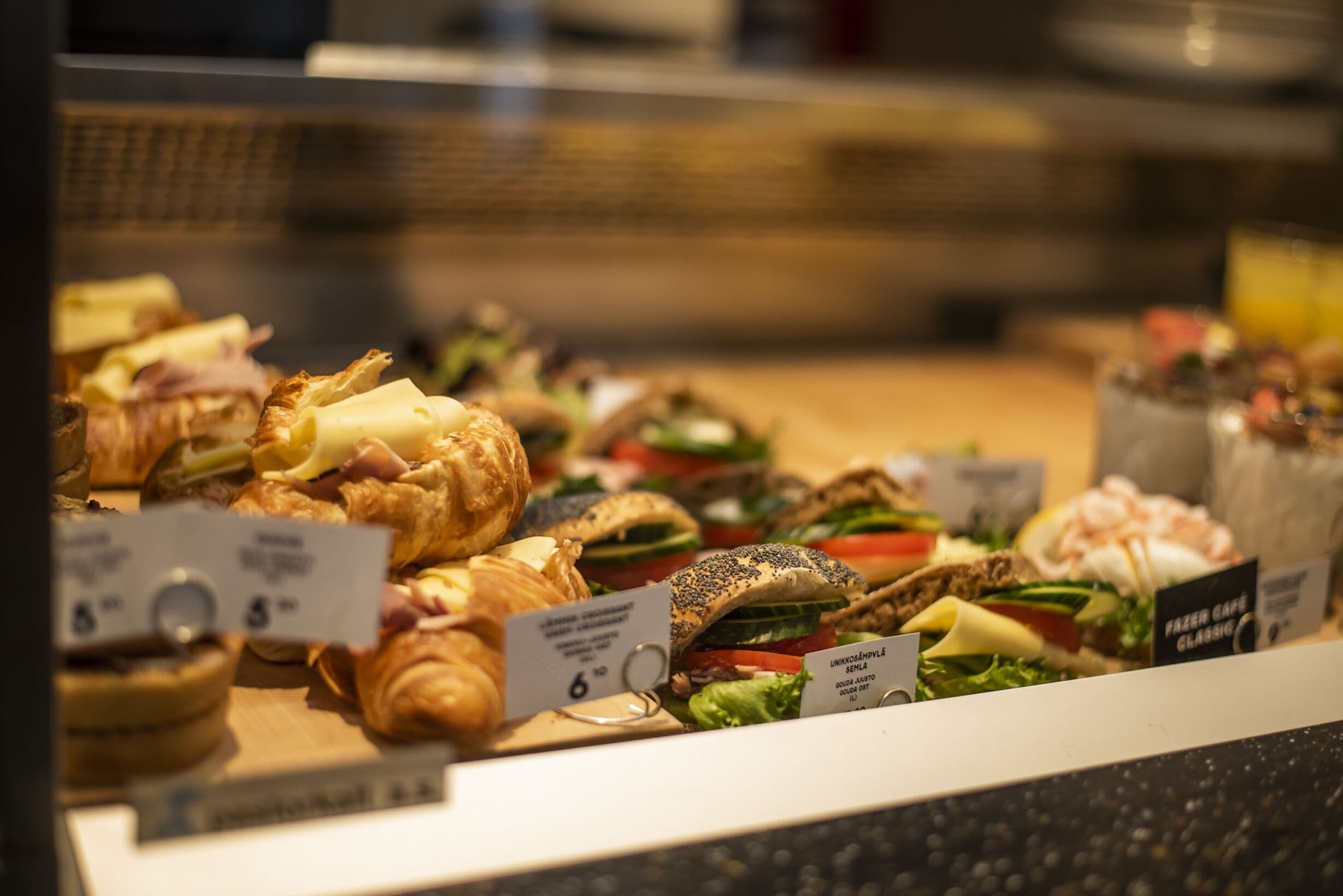 Variedad de sándwiches y pasteles expuestos en una vitrina con etiquetas de precios.