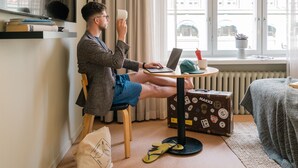 A man sitting at a desk with a laptop, a cup, and a suitcase in a room with a window, radiator, and a bed.