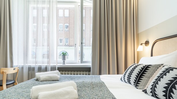 A hotel room with a bed, a chair, a window with curtains, and a view of buildings outside.