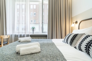 Habitación de hotel con cama, una silla, ventana con cortinas y vistas a edificios.