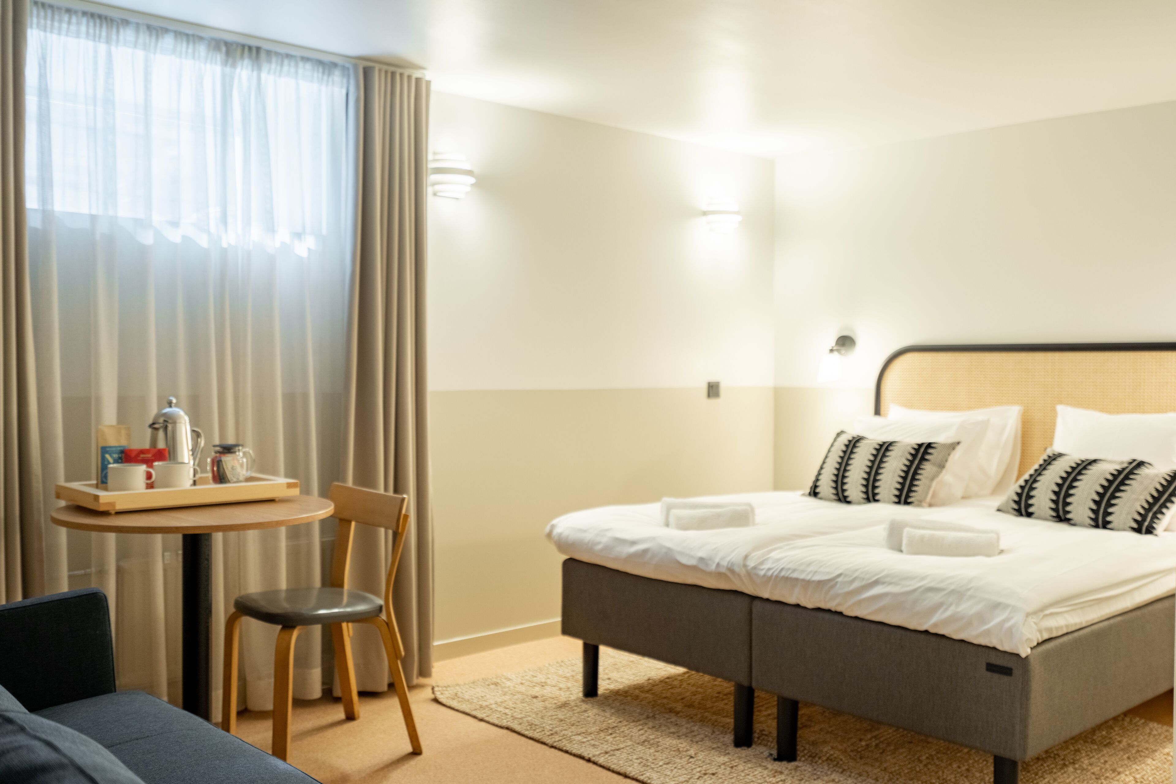 A hotel room with a bed, a small table with a tray of breakfast items, a chair, and a window with curtains.