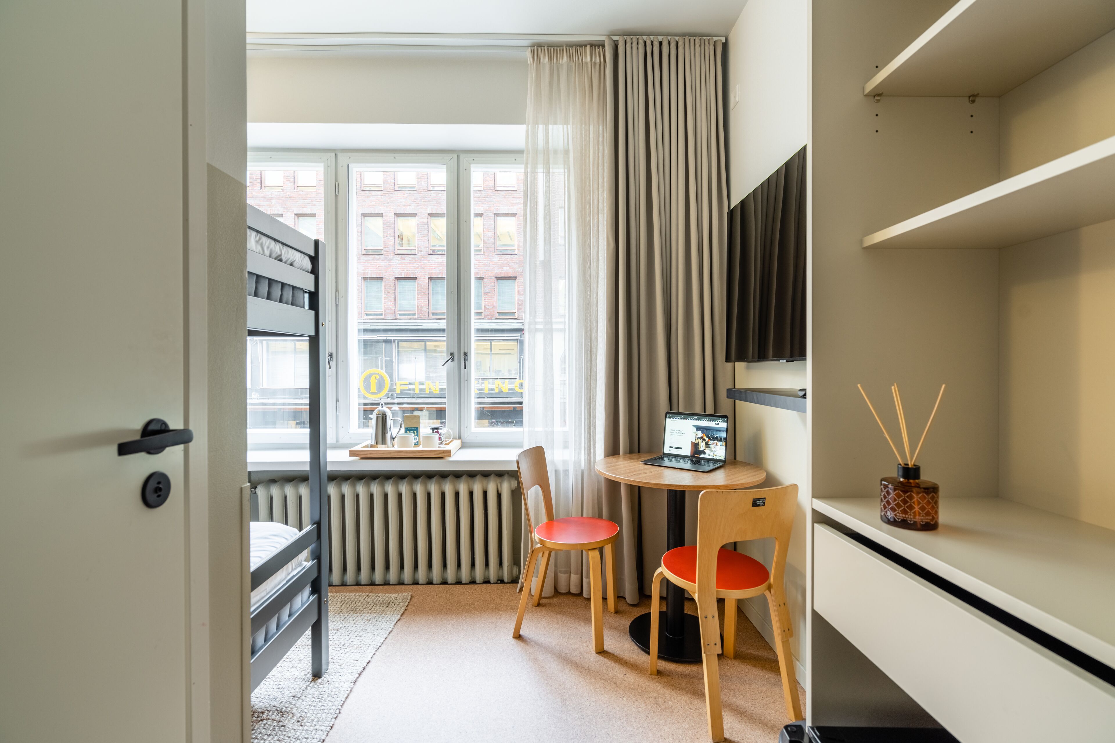 Une chambre d’hôtel de taille réduite, comprenant un lit, un bureau avec un ordinateur portable, deux chaises, un radiateur, une fenêtre avec des rideaux et une vue sur les bâtiments environnants.
