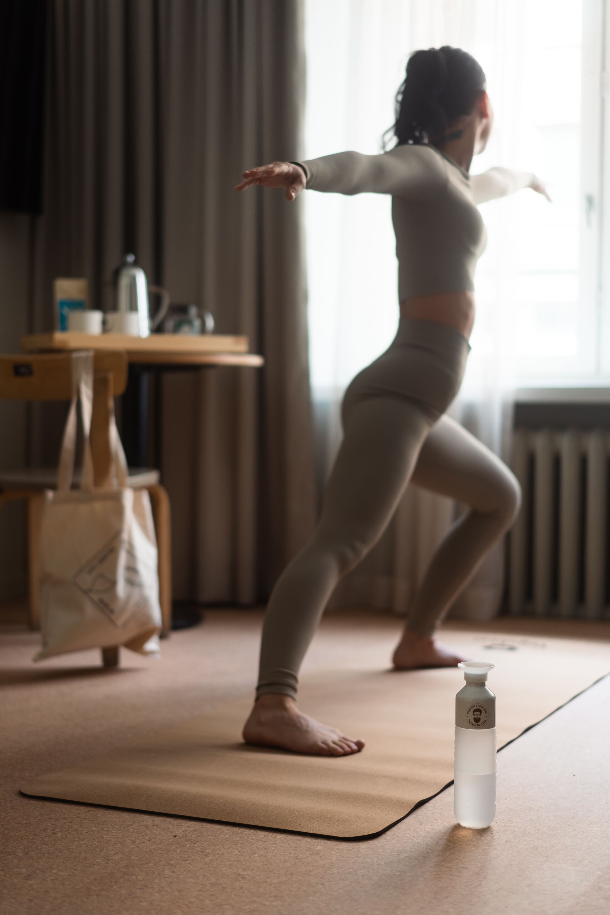 A person performing a yoga pose on a mat in a room with a radiator, a table with a bag, and a bottle on the floor.
