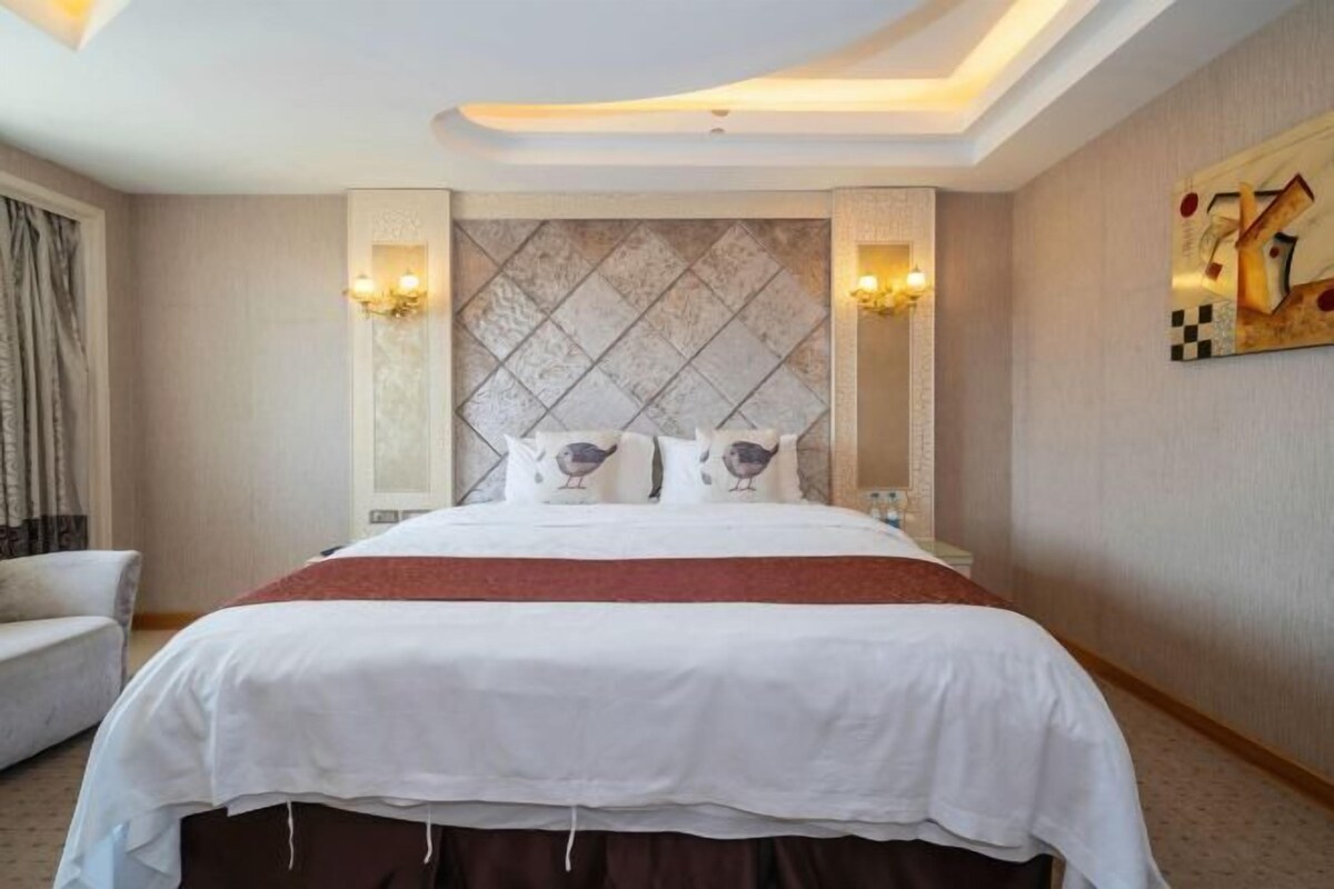 A hotel room with a large bed, a headboard with a diamond pattern, two wall-mounted lamps, and a painting on the wall.