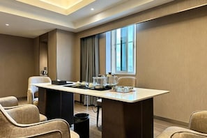 Un salón de conferencias de hotel con una larga mesa, varias sillas, una máquina de café y un ventanal con cortinas.