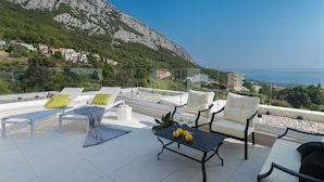 A rooftop terrace with white lounge chairs, a table with lemons, and a view of a mountainous coastline. - Villa Zvizdan (Makarska)