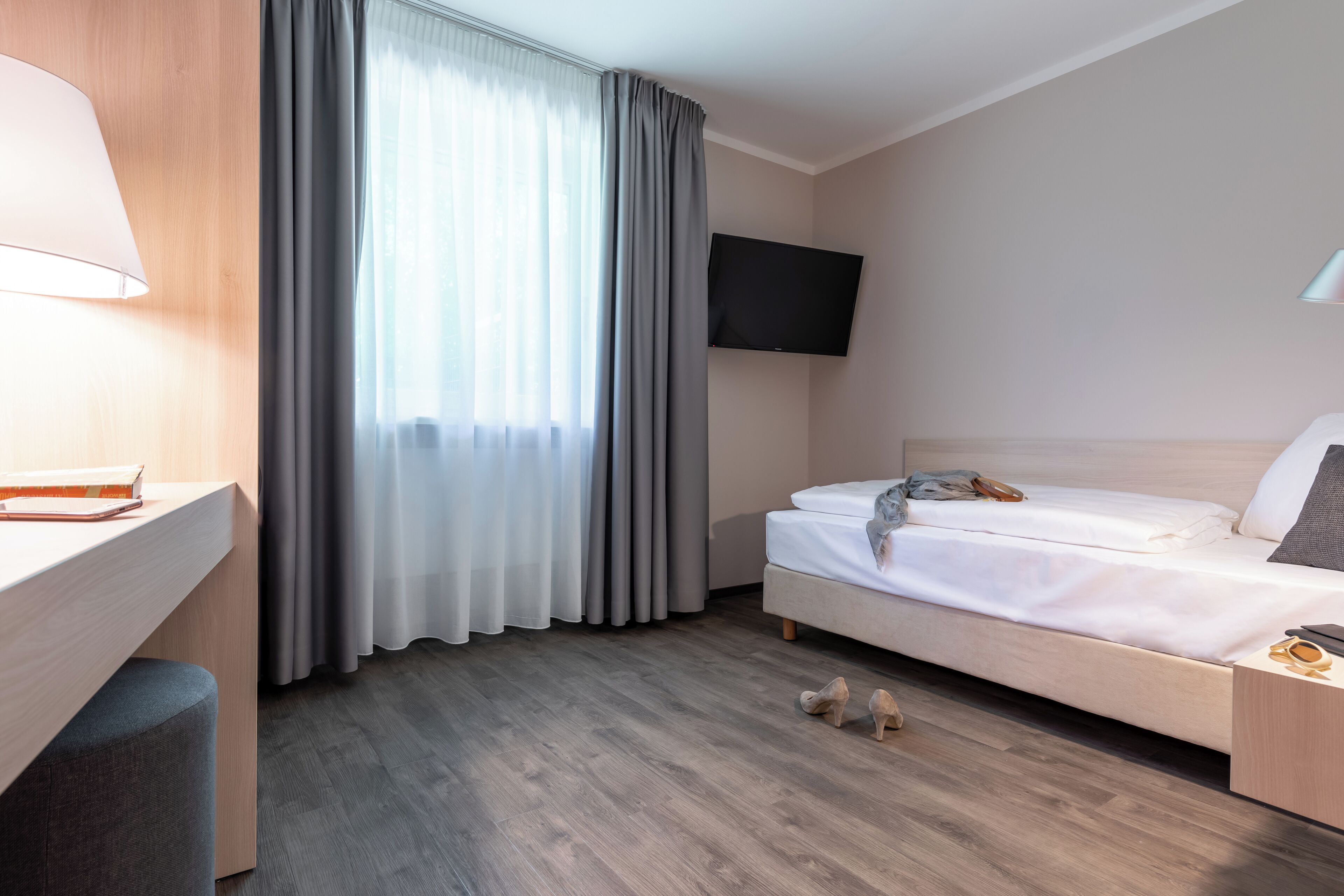 A hotel room with a wooden desk, a bed, a TV mounted on the wall, and a window with curtains.