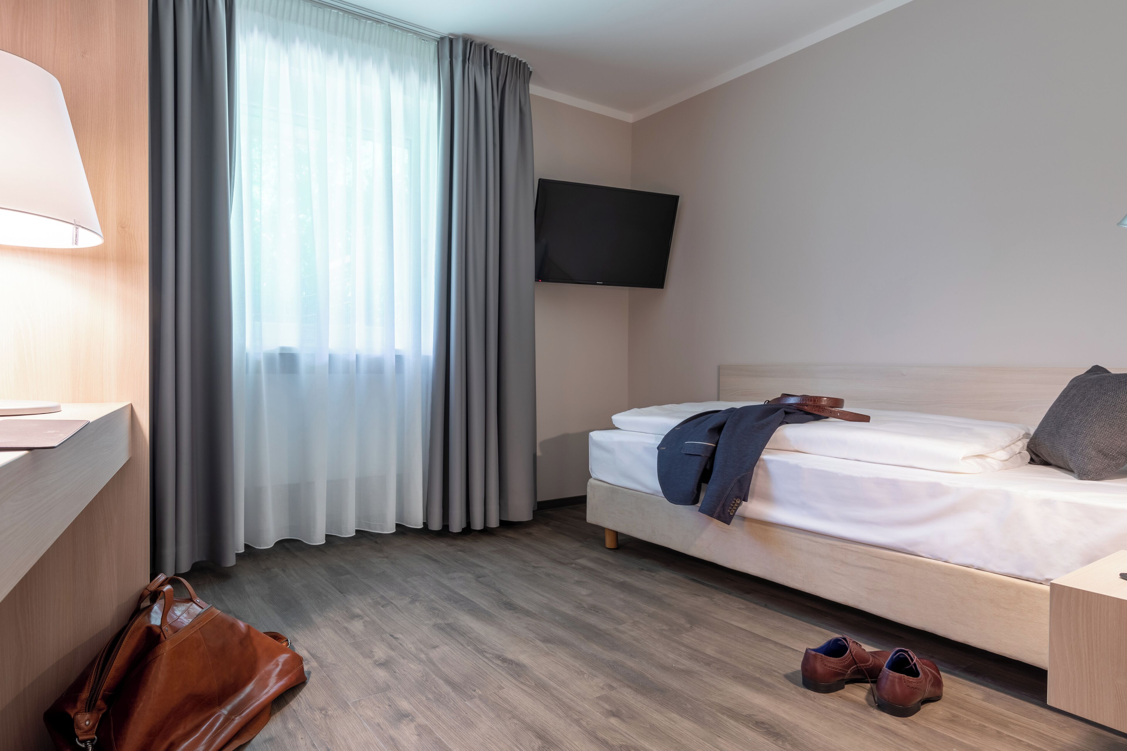Ein Hotelzimmer mit Bett, einem an der Wand befestigten Fernseher, einem Schreibtisch mit Lampe und einem Fenster mit Vorhängen.