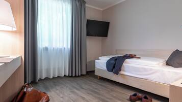 Ein Hotelzimmer mit Bett, einem an der Wand befestigten Fernseher, einem Schreibtisch mit Lampe und einem Fenster mit Vorhängen.