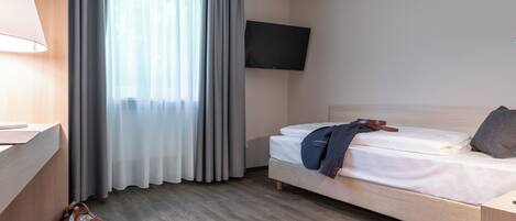 A hotel room with a bed, a TV mounted on the wall, a desk with a lamp, and a window with curtains.