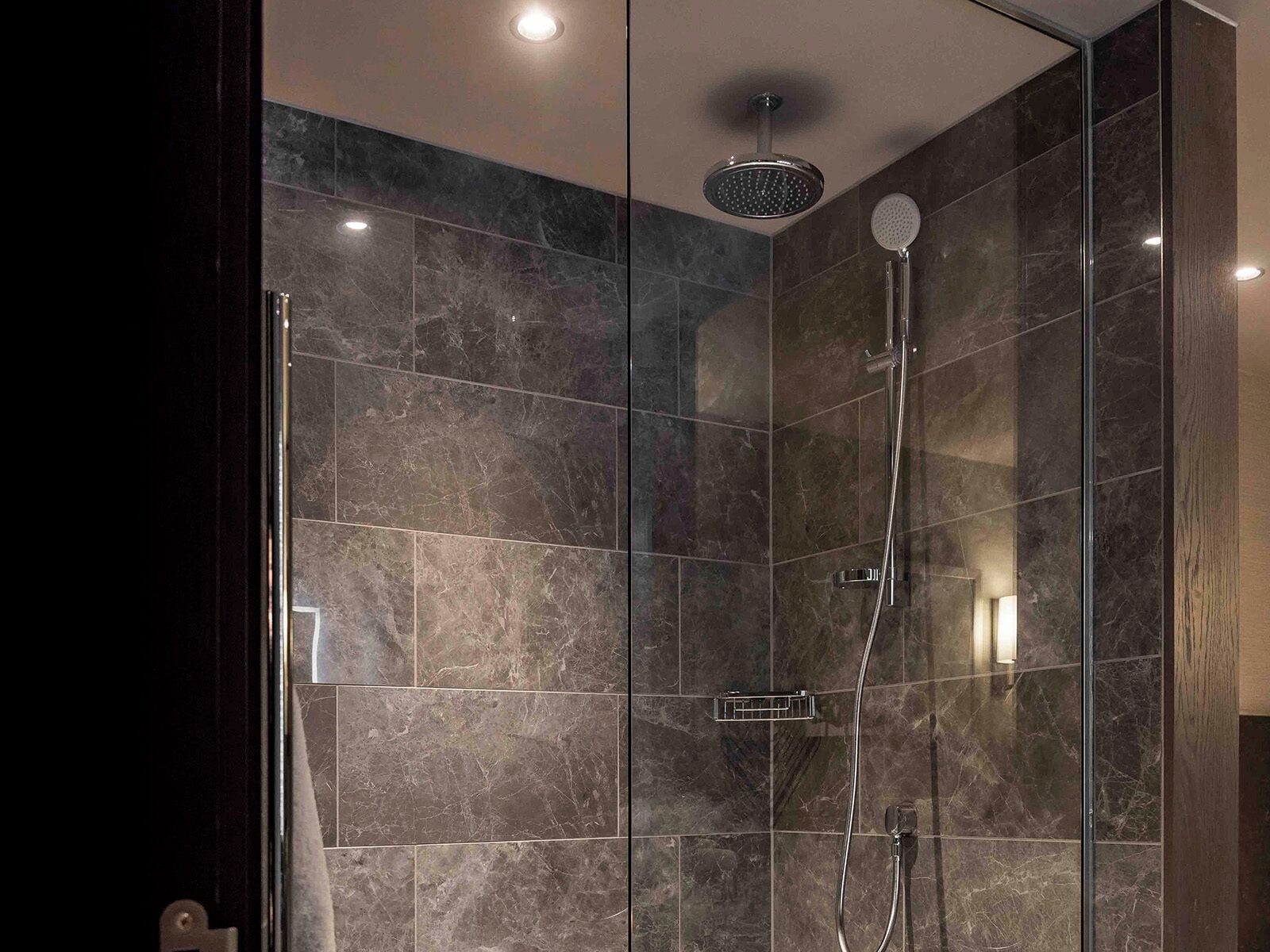 A modern shower with a handheld showerhead and a wall-mounted rain showerhead, surrounded by dark marble tiles.