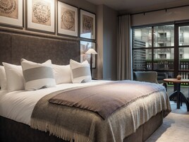 A modern hotel room with a large bed, a view of the city through floor-to-ceiling windows, and framed artwork on the wall.