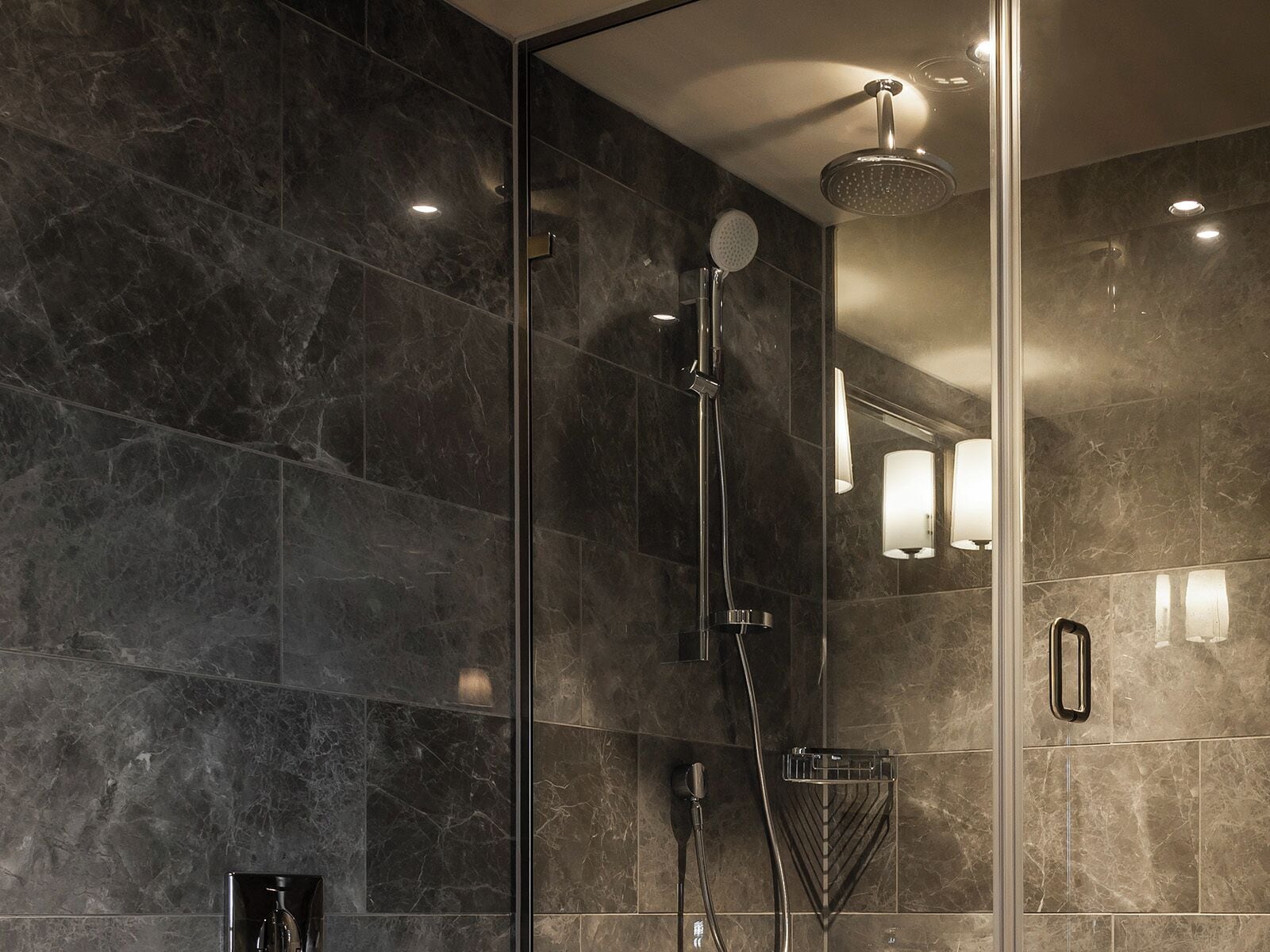 Une salle de bain moderne avec des carreaux de marbre foncé, une douche en verre avec un pommeau de douche à main et un luminaire fixé au mur.